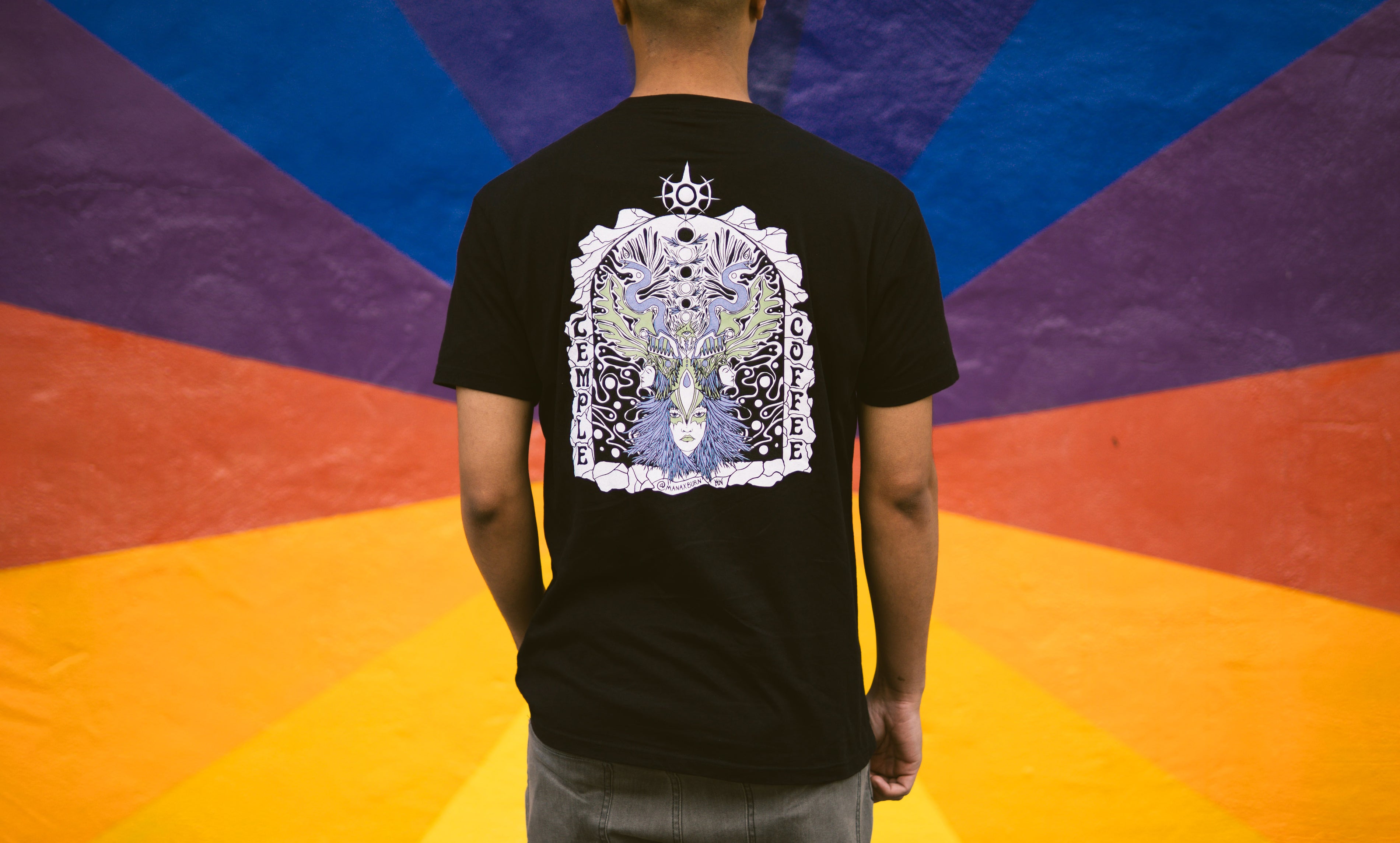 Man standing in front of colorful mural with a black t-shirt with an intricate print design.
