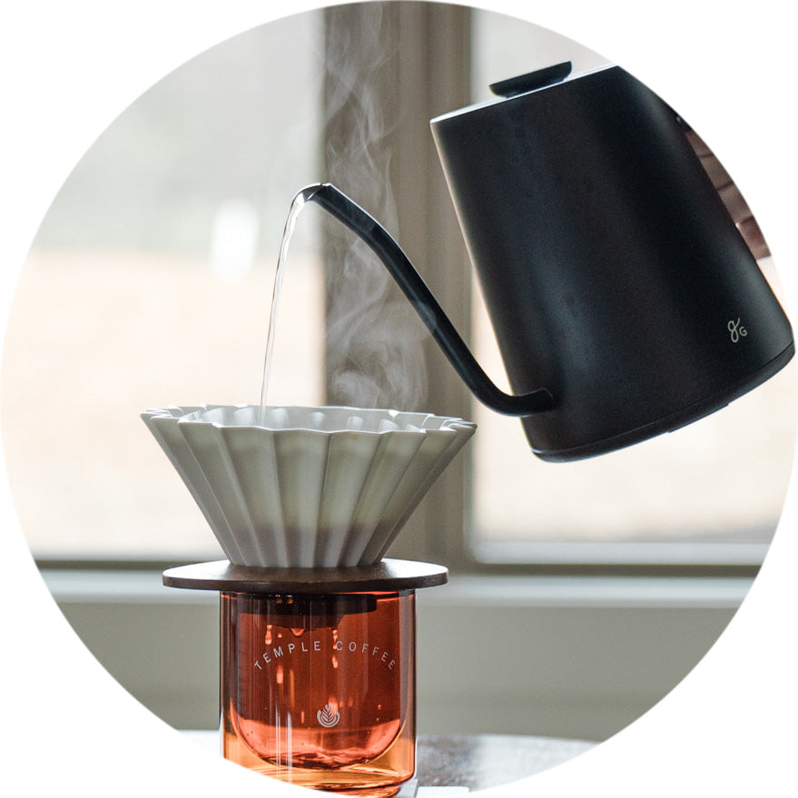 Black kettle pouring water into a ceramic coffee dripper with a glass container below, on a neutral background.