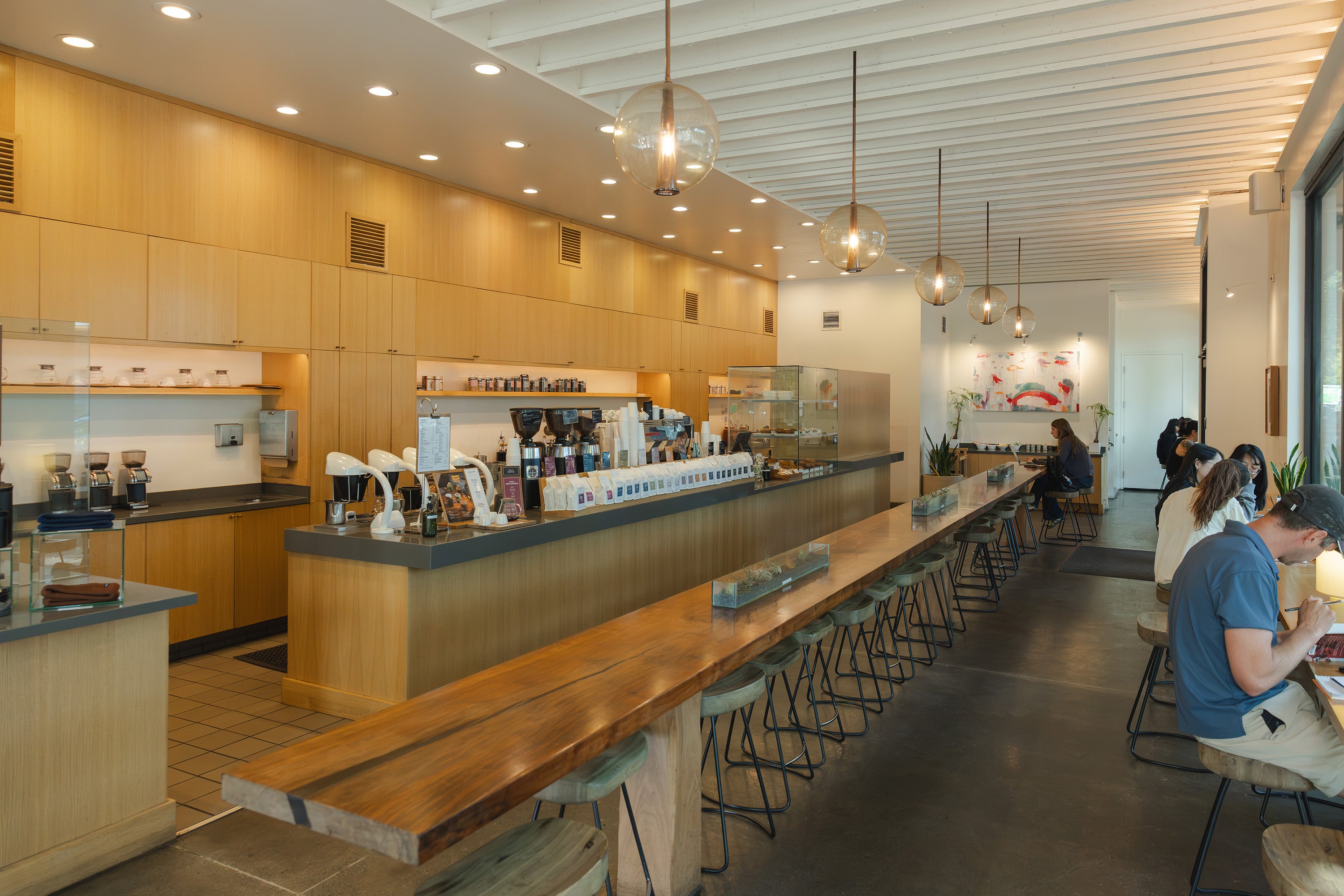 A modern coffee shop interior with a long wooden counter, bar stools, hanging pendant lights, and several people seated and working near the counter.