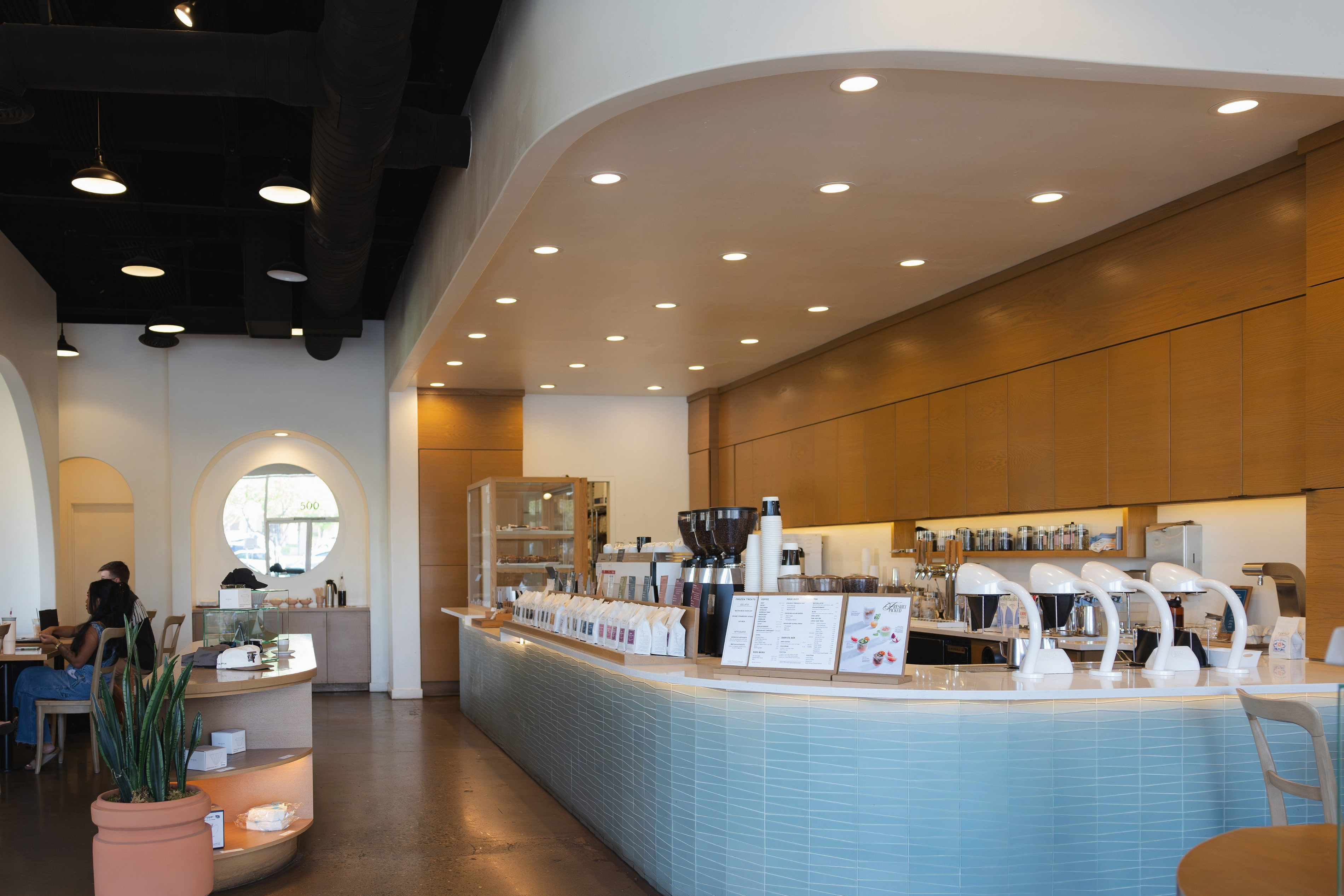A modern coffee shop interior with a long light blue tiled counter, espresso machines, warm wood wall panels, recessed ceiling lights, and a few seated people in the background.