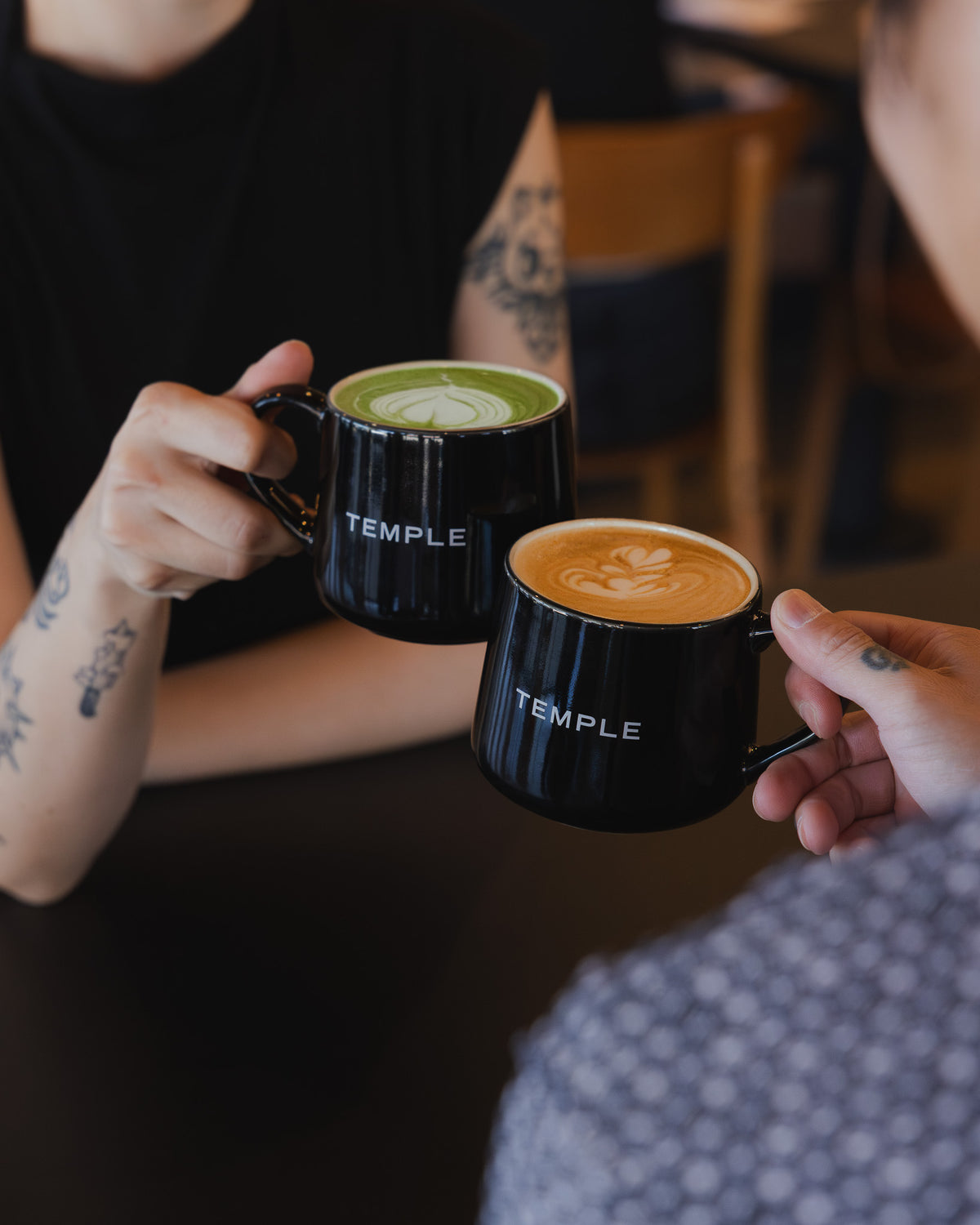 Two people holding black mugs with 'Temple' branding, one containing a green drink and the other a coffee drink.