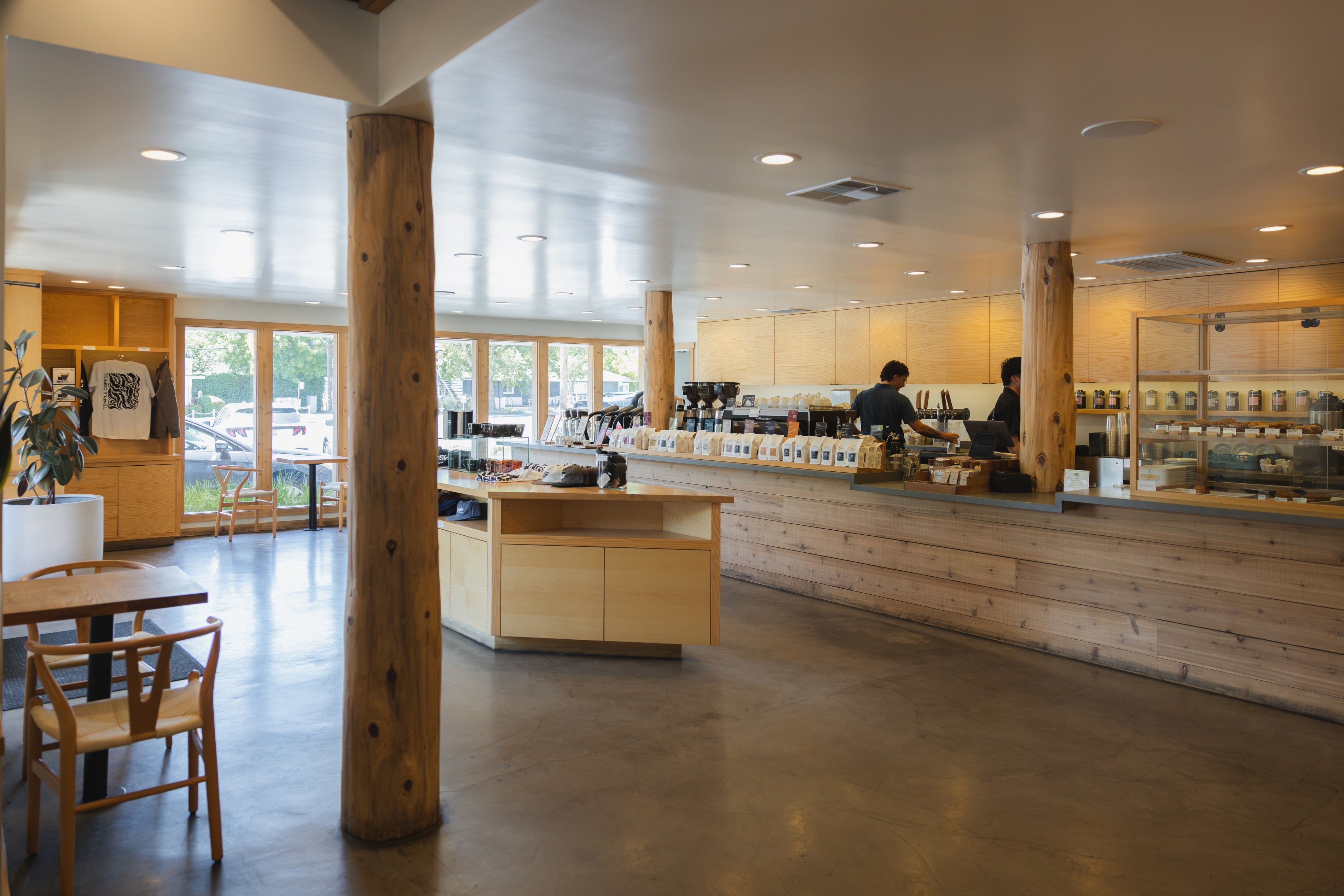 A spacious, modern cafe interior with a long wooden counter, wood pillars, concrete floors, and large windows in the background. Two staff members are visible behind the counter.