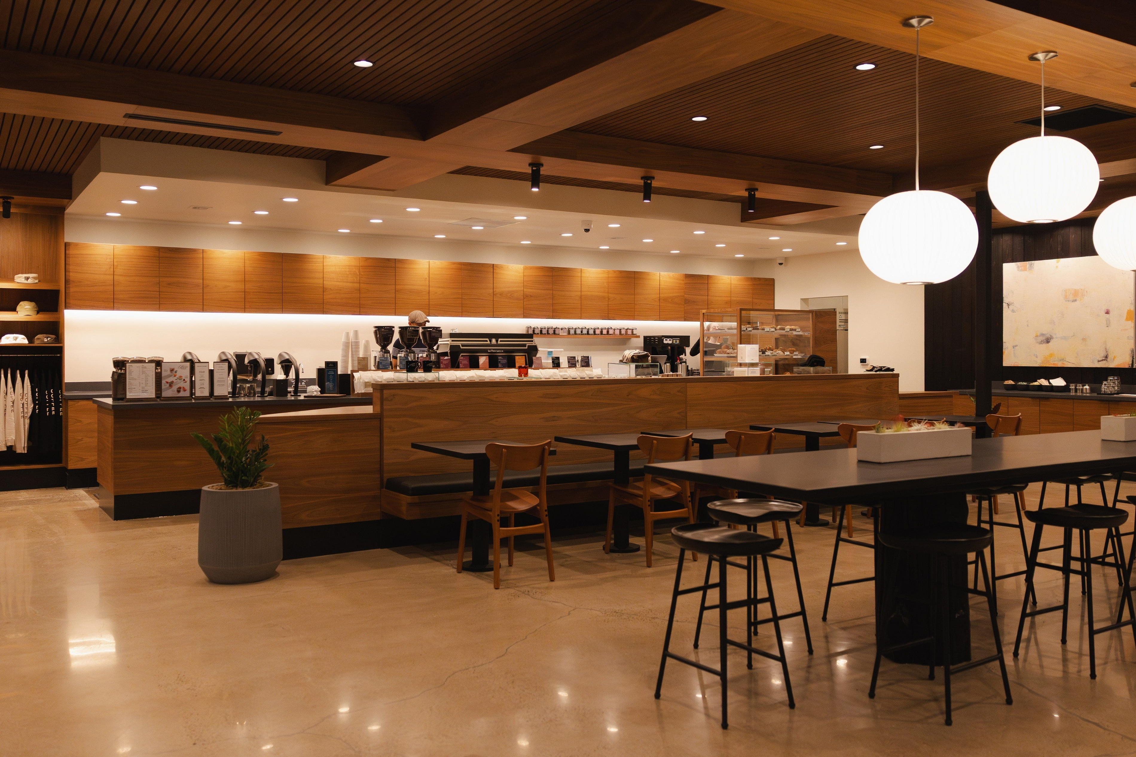 A modern cafe interior with a wood counter, bar stools, a high black prep table, pendant lights, and warm wood ceiling panels.