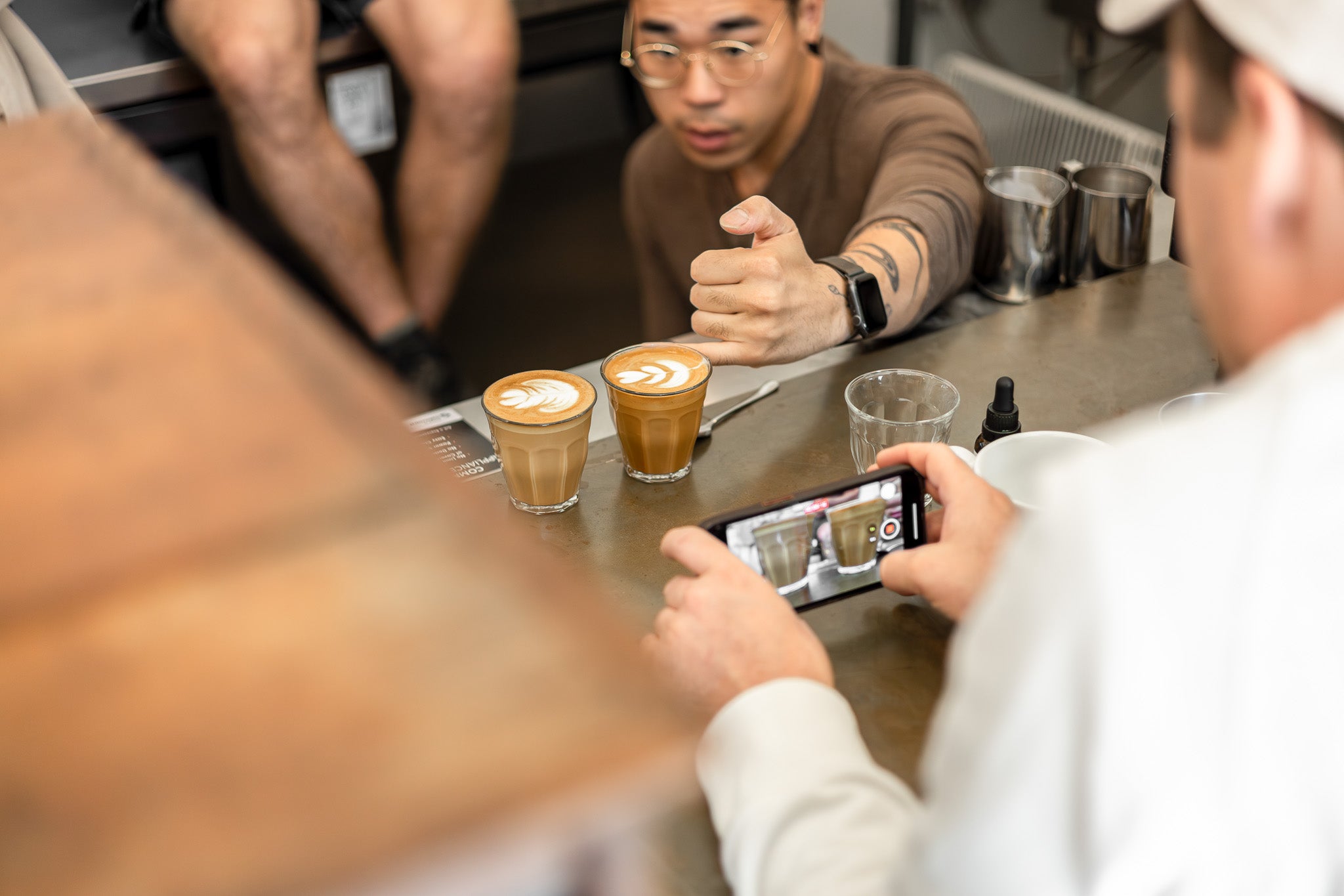 Two people at a table with coffee drinks, one taking a photo with a smartphone.