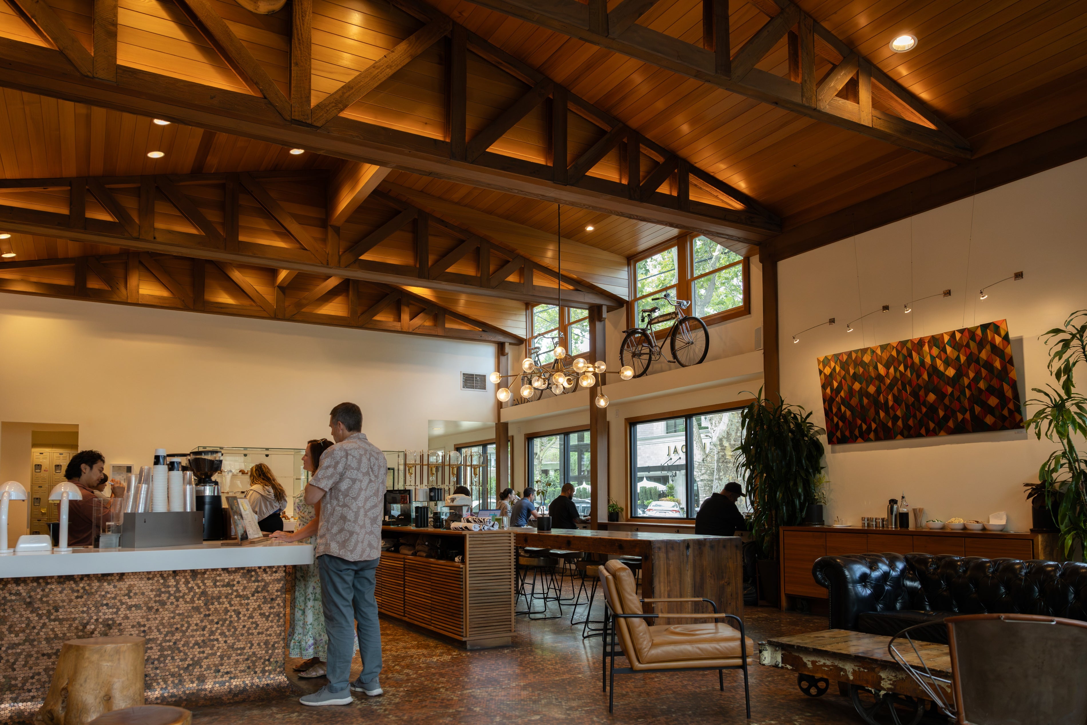 A wide interior shot of a rustic cafe or coffee shop with a high wood-beam ceiling, a counter with baristas, seated customers, chairs, plants, and a large textured wall art piece.